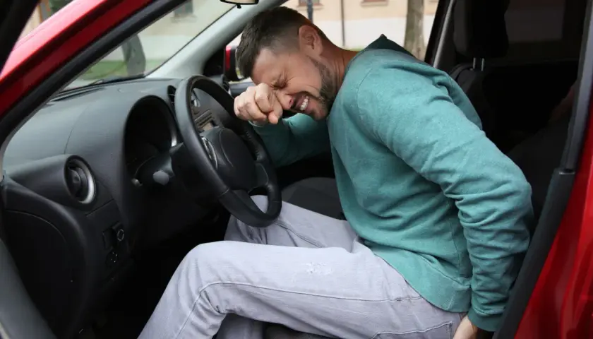 Mężczyzna w samochodzie z bólem pośladka, trzymający się za bolące miejsce.