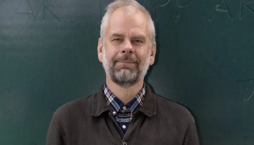 Anders, med grått hår och skägg, ler mot kameran framför en grön tavla. Han bär en rutig skjorta under en mörk jacka.