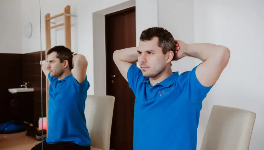 Mężczyzna w niebieskiej koszulce wykonuje ćwiczenia na szyję, opierając dłonie na karku i delikatnie napinając mięśnie.