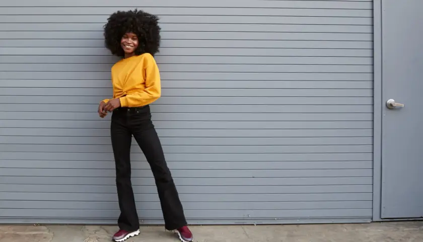 Une jeune femme souriante pose devant un mur gris, portant un haut jaune et un jean flare. Quelles chaussures avec jean flare ? Des baskets bordeaux complètent ce look décontracté.