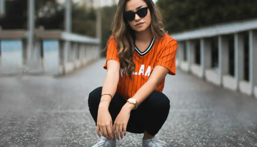 Chica con gafas de sol y camiseta naranja a rayas, agachada en un puente. Su atuendo es muy comfy y moderno.