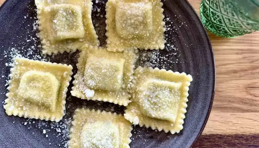 Pięć kwadratowych ravioli z ricottą, posypanych parmezanem, na ciemnym talerzu. Obok zielony kieliszek.