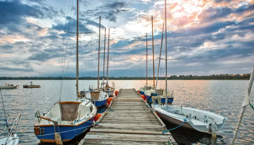 Słoneczny zachód nad pogorią jezioro. Drewniane molo z żaglówkami, chmury odbijające się w wodzie.