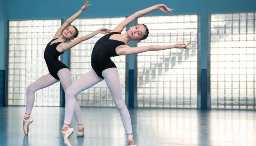 Dwie młode tancerki baletowe w czarnych trykotach i białych rajstopach, stojące na czubkach palców, wykonują elegancką choreografię.
