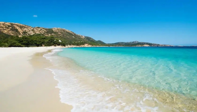 Plage de sable blanc bordée par une eau turquoise cristalline, avec des collines verdoyantes en arrière-plan. Un paysage idyllique d'**Italie bord de mer**.