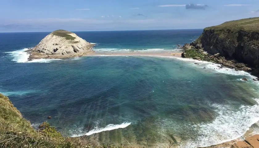 Vista de la Virgen del Mar en Santander, con una isla rocosa y aguas turquesas.