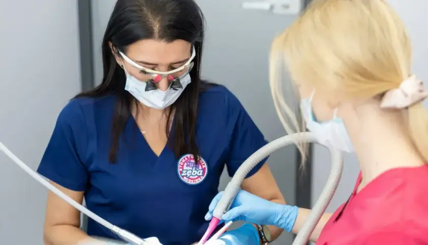 Dwie dentystki w maseczkach i okularach pracują przy pacjencie. Jedna używa wiertła, druga odsysacza. To profesjonalna artykaina.