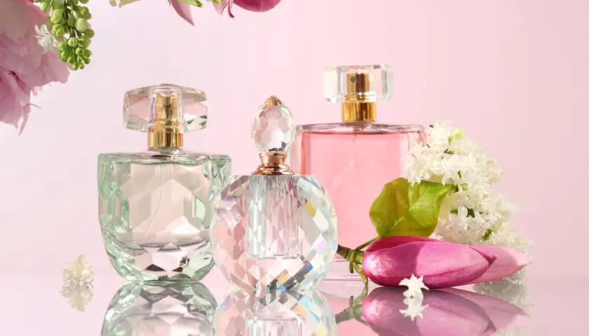 Trzy flakoniki perfum i kwiaty na różowym tle.