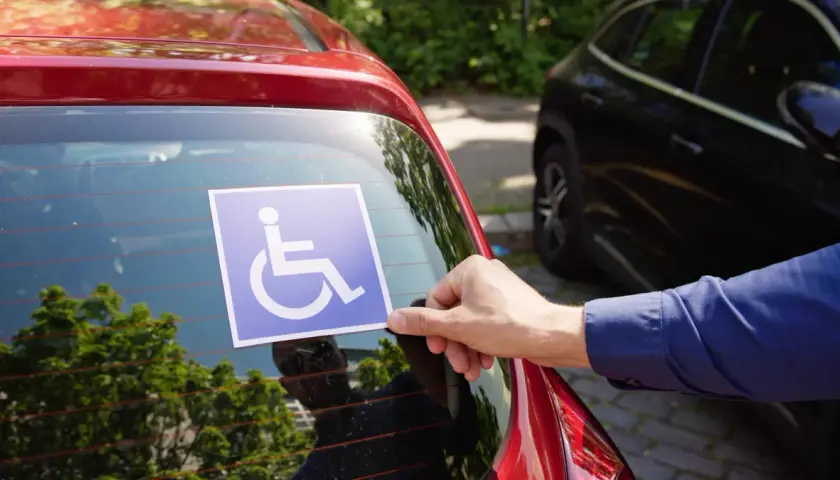 Ręka przykleja niebieską naklejkę z symbolem osoby na wózku inwalidzkim na tylnej szybie czerwonego samochodu. To przypomnienie o tym, jakie dokumenty do karty parkingowej są potrzebne.