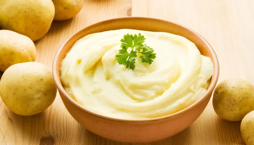 Un tazón de cremoso puré de patatas, decorado con perejil, junto a patatas crudas. ¡Así es como hacer puré de patatas perfecto!