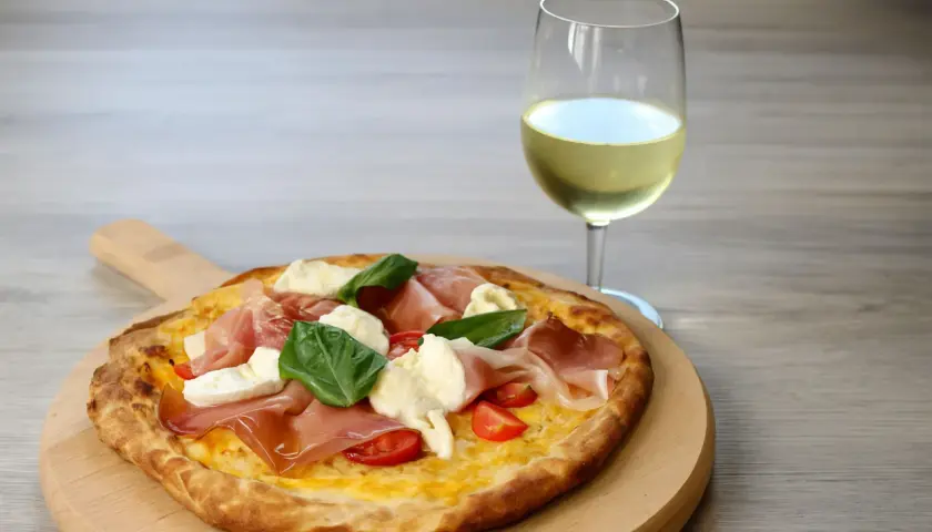 Pizza con prosciutto, mozzarella e basilico su tagliere di legno, accanto a un bicchiere di vino bianco. Un perfetto abbinamento vino pizza.