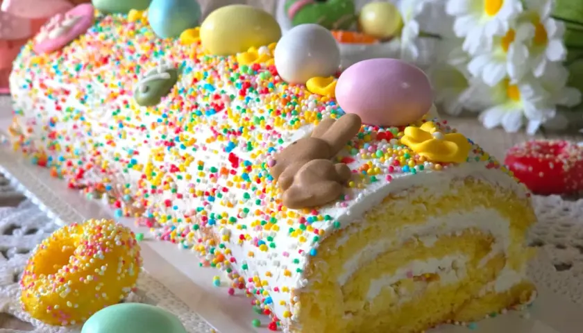 Bûche roulée décorée pour Pâques, recouverte d'un glaçage mascarpone facile et de vermicelles colorés, avec des œufs et un lapin en chocolat.