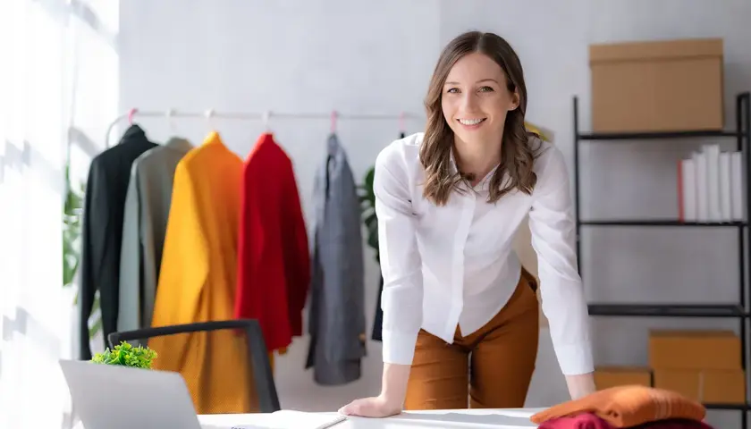 Une chef de produit textile souriante, entourée de vêtements colorés sur un portant, travaille dans son bureau lumineux.