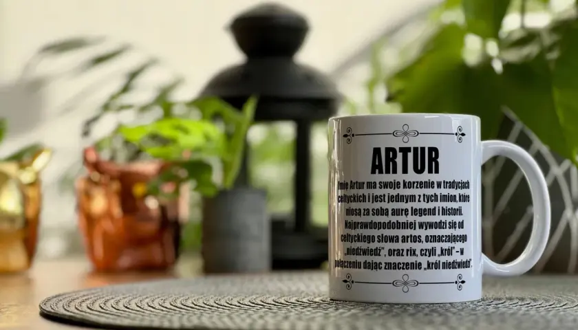 Biały kubek z napisem "ARTUR" i tekstem o znaczeniu imienia, na tle roślin.
