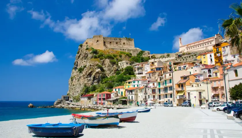 Vue d'une ville côtière italienne avec un château sur une falaise, des maisons colorées et des bateaux sur une plage.