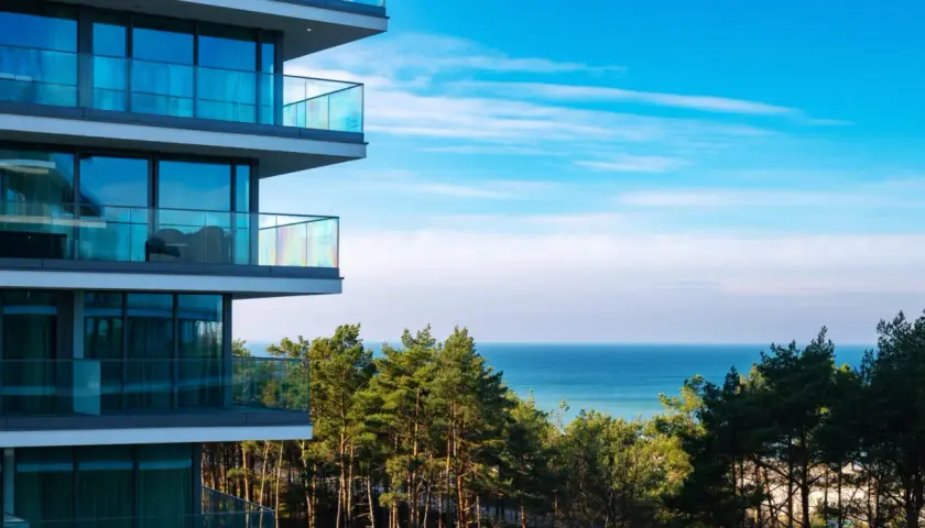Nowoczesny budynek z balkonami i widokiem na morze. W oddali las i błękitne niebo. Idealne miejsce na wypoczynek w Wave Międzyzdroje Resort&SPA.