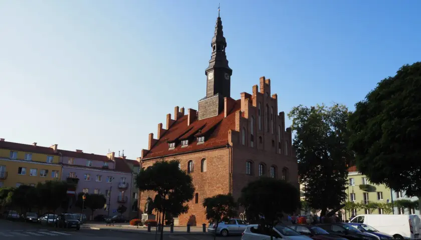 Ratusz w mieście nad jeziorem Skiertąg, z charakterystyczną wieżą zegarową, otoczony kolorowymi kamienicami i zielenią.