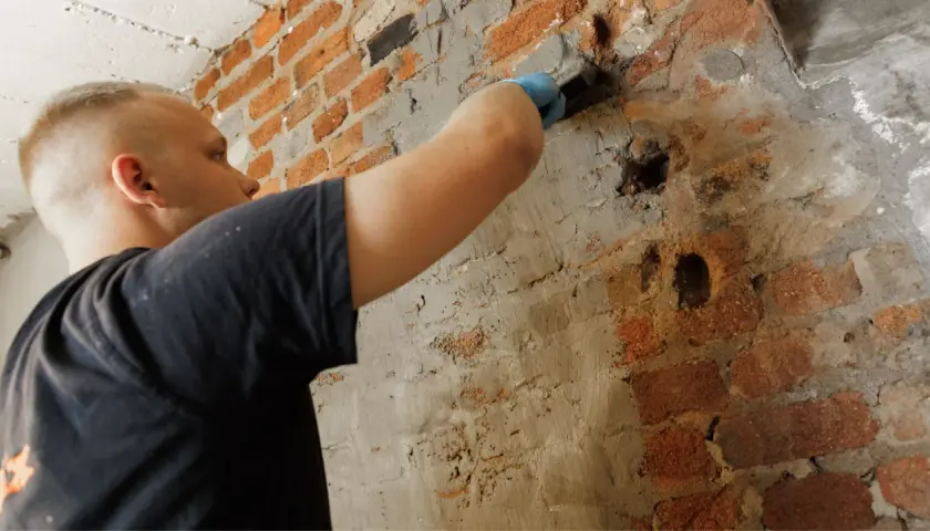 Mann mit blauer Handschuh verputzt eine alte Ziegelwand. Die **Bitumen Hauswand** wird repariert.