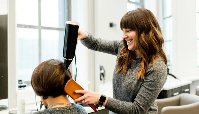 Estilista sonríe mientras seca el cabello de una clienta. Un buen momento para cortarse el pelo es cuando te sientes renovada.