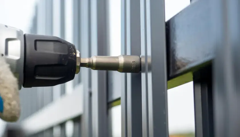Montaż elektrozaczepu do furtki. Wiertarka z nasadką 8mm przykręca element do metalowej konstrukcji.
