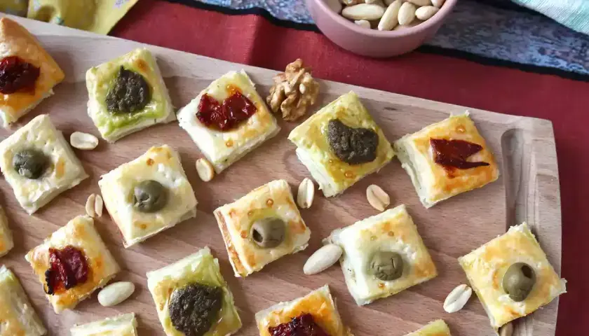 Rustici con pasta sfoglia, quadrati dorati con olive, pesto e pomodori secchi, sparsi su un tagliere con frutta secca.