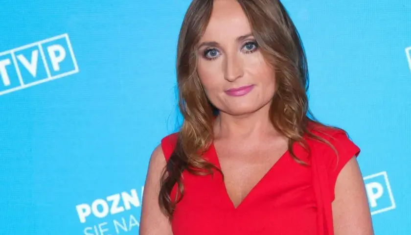 Justyna Dobrosz-Oracz w czerwonej sukience, na tle niebieskiej ściany z logo TVP.