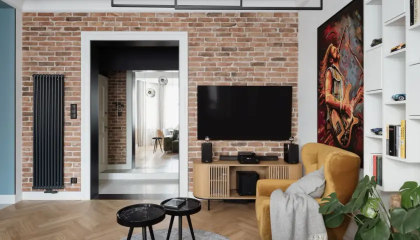 Nowoczesny salon z ceglaną ścianą, na której wisi telewizor. Oświetlenie ściany z kamienia podkreśla jej fakturę. Obok żółty fotel i obraz z gitarzystą.