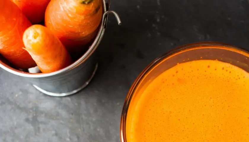 Frische Karotten in einem Eimer neben einem Glas leuchtend orangem Karottensaft. Die Wirkung von Karottensaft ist bekannt für seine Vitamine.