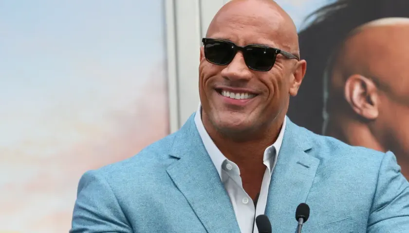 Dwayne "The Rock" Johnson, być może najbogatszy aktor na świecie, w błękitnej marynarce i okularach przeciwsłonecznych, uśmiecha się do kamery.