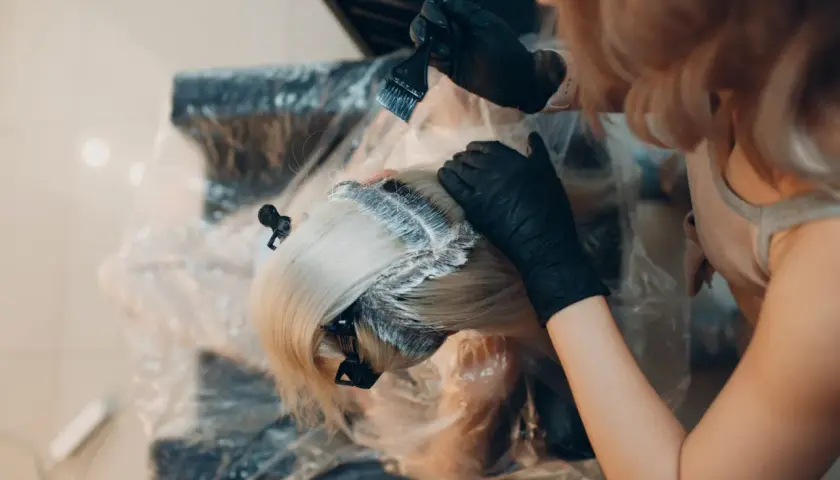 Fryzjerka w czarnych rękawiczkach nakłada farbę na blond włosy. Kluczowe jest, ile trzymać farbę na włosach, by uzyskać pożądany efekt.