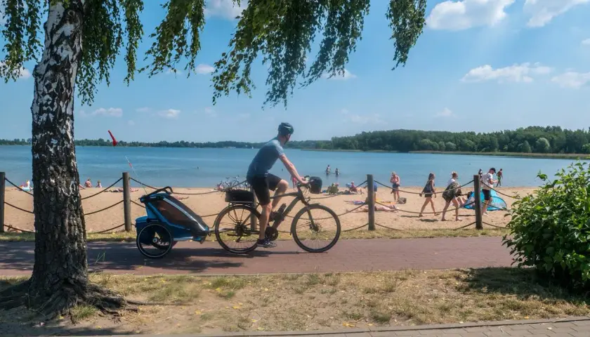 Rowerzysta z przyczepką na plaży nad jeziorem, w tle ludzie odpoczywający na piasku. Trasa Pogoria 3.