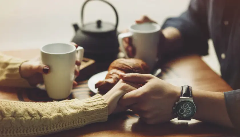 Dłonie trzymające kubki z kawą, croissant na talerzyku. W tle czajniczek. To idealny moment na szczere komplementy dla kobiety.