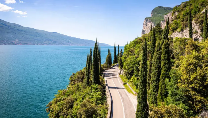 Route panoramique longeant un lac italien, idéale pour le camping-car. Cyprès élancés, falaises et eau bleue invitent à l'aventure.