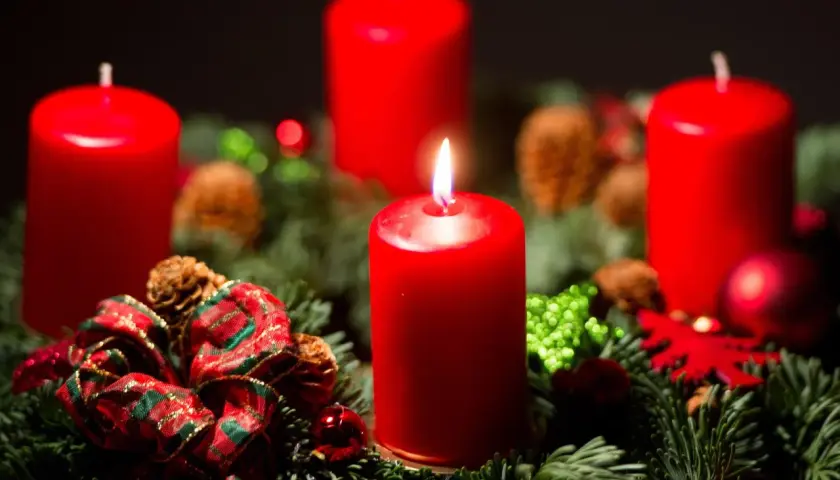 Ein Adventskranz mit vier roten Kerzen, Tannenzweigen, Zapfen und Schleifen. Die erste Kerze brennt und symbolisiert, warum man Advent feiert: die Vorfreude auf Weihnachten.