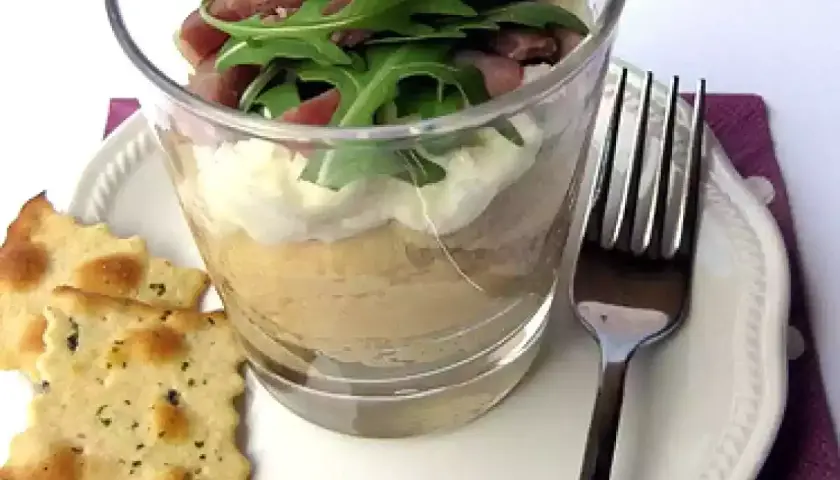 Deliziosa crema di ceci in bicchiere, stratificata con formaggio spalmabile e guarnita con rucola e prosciutto. Servita con cracker croccanti.