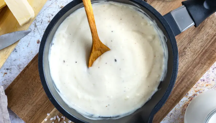 Cremige Bechamel-Sauce in einer Pfanne, bereit für die Lasagne. Butter, Milch und Gewürze sind daneben zu sehen.