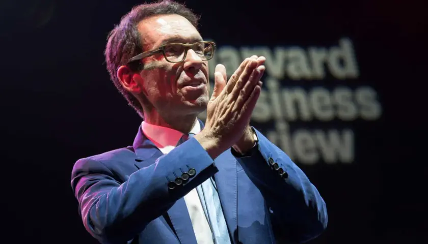 Byron Sharp, w okularach i niebieskim garniturze, z dłońmi złożonymi w geście przemowy, na tle napisu "Harvard Business Review".