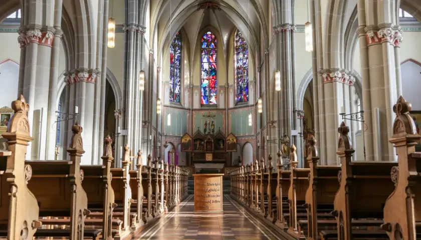 Blick in den katholischen Kircheninnenraum mit Holzbänken, Buntglasfenstern und einem Altar.
