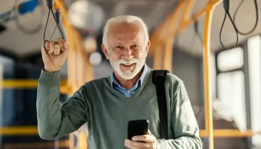 Uśmiechnięty starszy pan z telefonem w dłoni, trzymający się poręczy w pojeździe. Cieszy się z możliwości 70 lat darmowe przejazdy PKP Intercity.