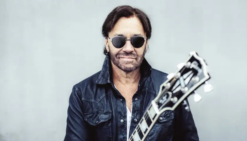 Al Di Meola w czarnej skórzanej kurtce i okularach przeciwsłonecznych, trzymający gitarę.