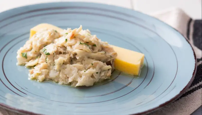 Baccalà alla Vicentina, ein cremiges Stockfischgericht mit Petersilie, serviert mit einem Stück Polenta auf einem blauen Teller.