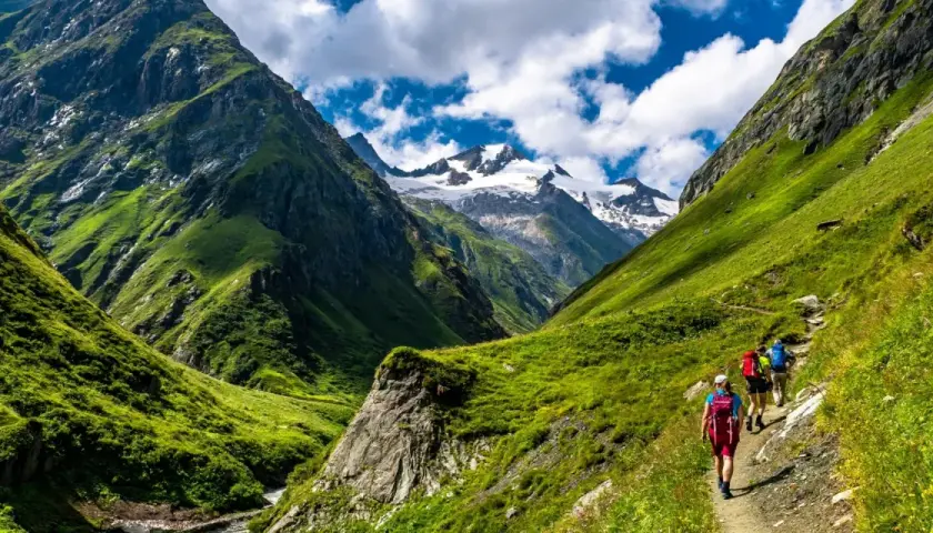 Hikers on a trail enjoying alpine hiking amidst lush green mountains and snow-capped peaks under a cloudy blue sky.