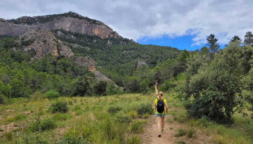 Senderista camina por un sendero en los **estrets d'Arnes**, rodeada de pinos y montañas rocosas bajo un cielo nublado.