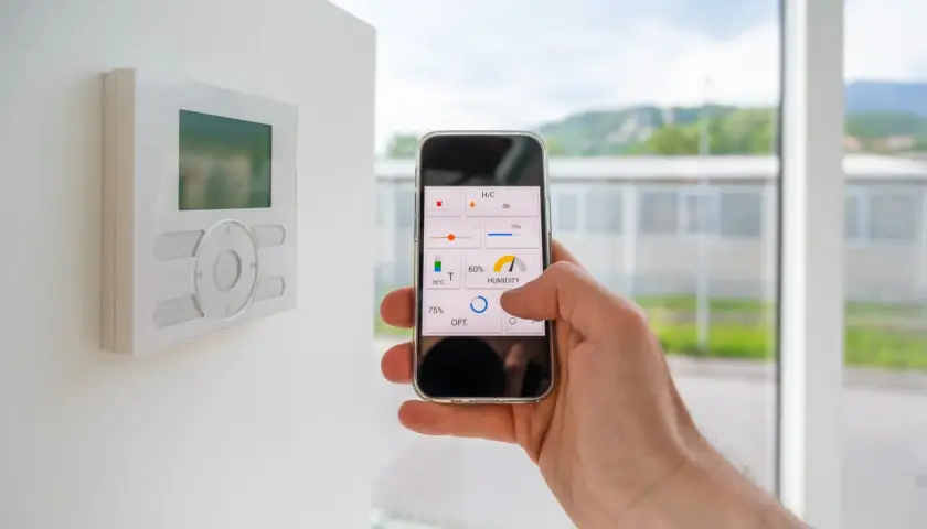 Control de la automatización de casas desde un smartphone. Se ve un termostato en la pared y una mano sujetando un teléfono con una app de control.