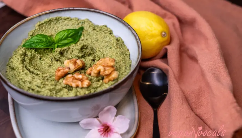 Un delicioso pesto vegano en un cuenco, adornado con hojas de albahaca y nueces. Junto a un limón y una flor rosa.