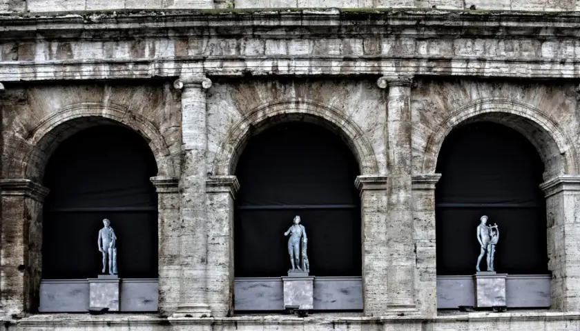 Tre statue classiche emergono dall'ombra di un imponente arco a Roma, testimoni silenziose di un passato glorioso.