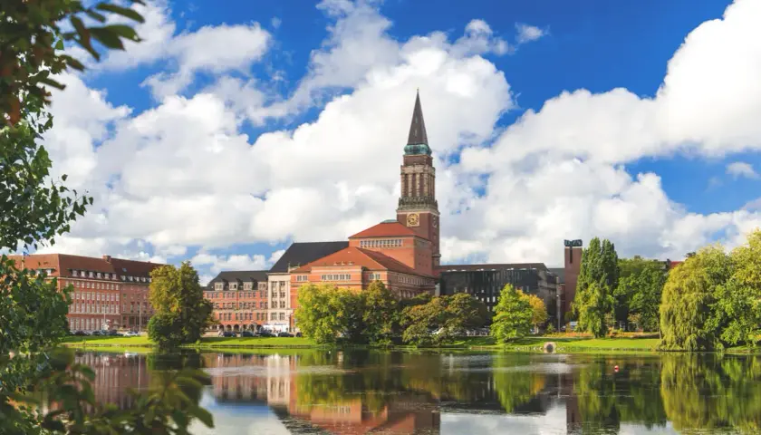 Ein malerischer Blick auf das Rathaus von Kiel, perfekt für Kiel Sightseeing. Der Turm ragt in den blauen Himmel, während sich das Gebäude im ruhigen Wasser spiegelt.