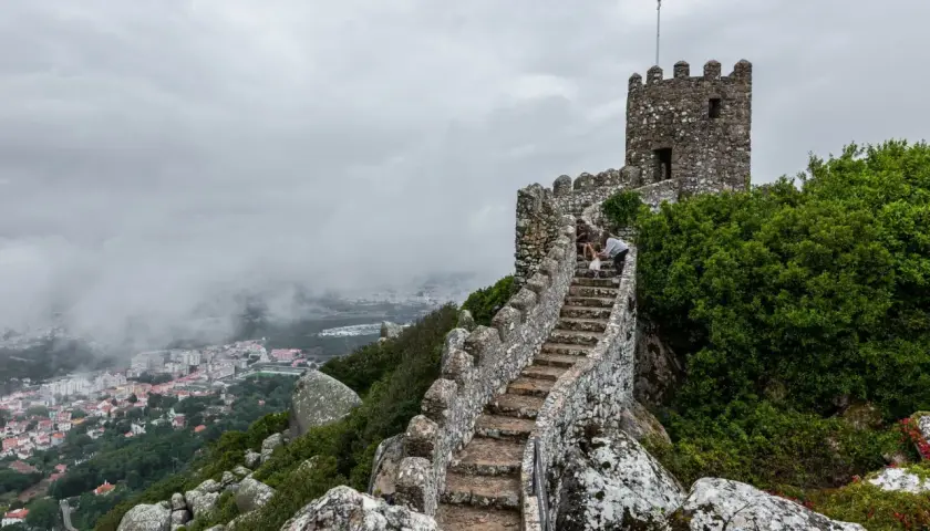 Escaleras de piedra ascienden hacia una torre en el **castillo de Sintra**, con vistas a la ciudad envuelta en niebla.