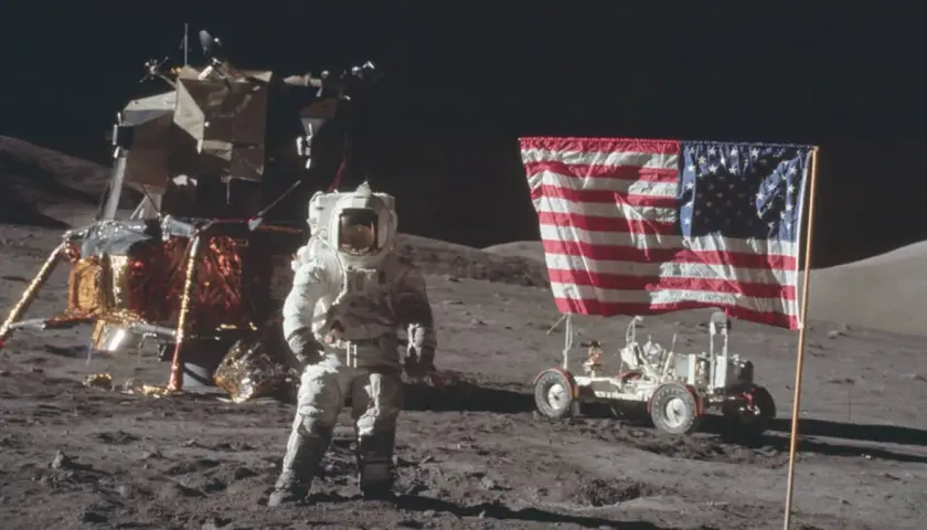 Astronaut w skafandrze podczas spaceru po księżycu, obok lądownika i łazika z amerykańską flagą.