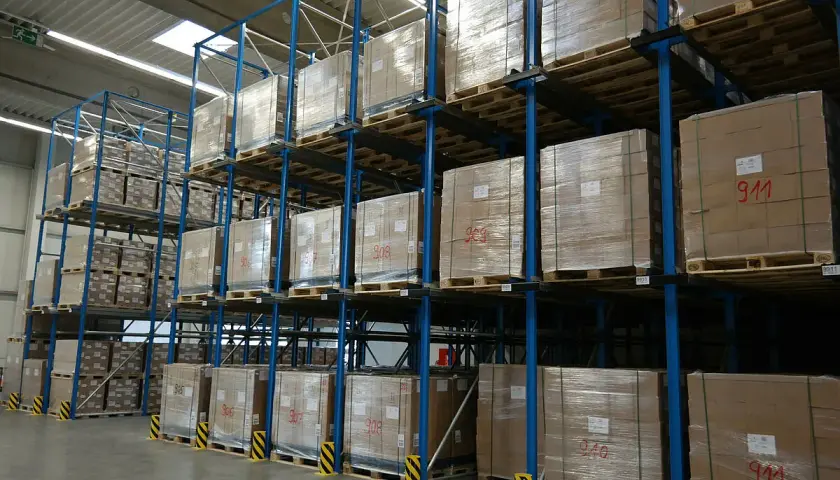 Wysoki magazyn centralny z regałami pełnymi paczek na paletach. Widać oznaczenia na kartonach, np. "911".